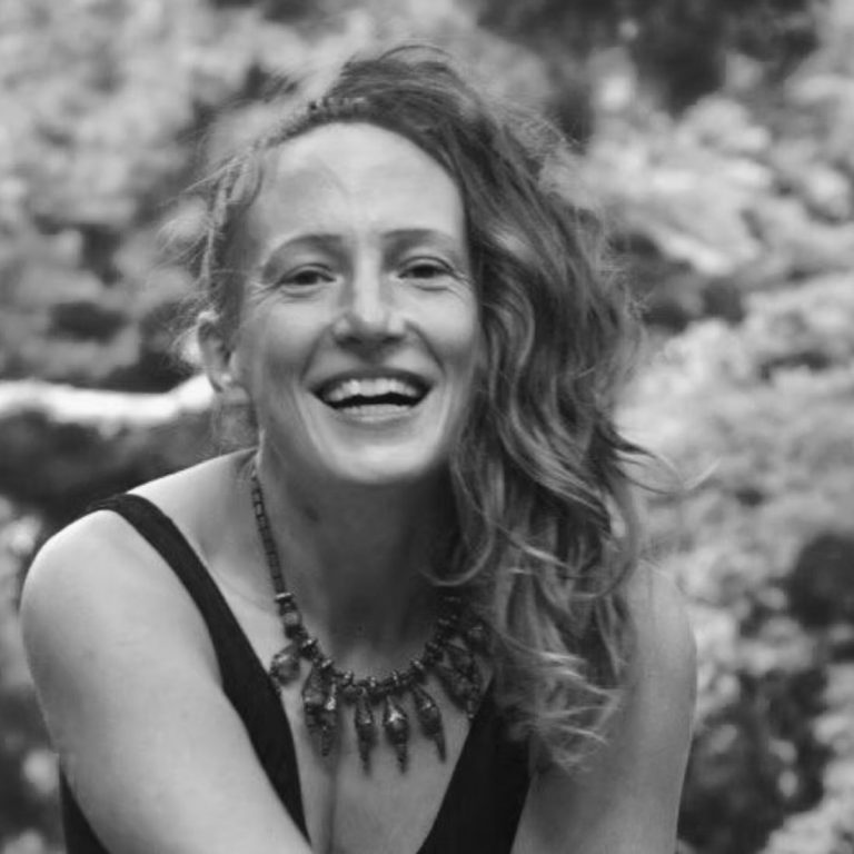 Jennifer Nausch Lächelnde Frau mit lockigem Haar, trägt eine Halskette, Hintergrund mit natürlicher Vegetation.