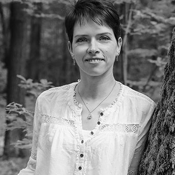 Jenny Tausendfreund Frau steht lächelnd an einem Baum im Wald, trägt eine weiße Bluse.