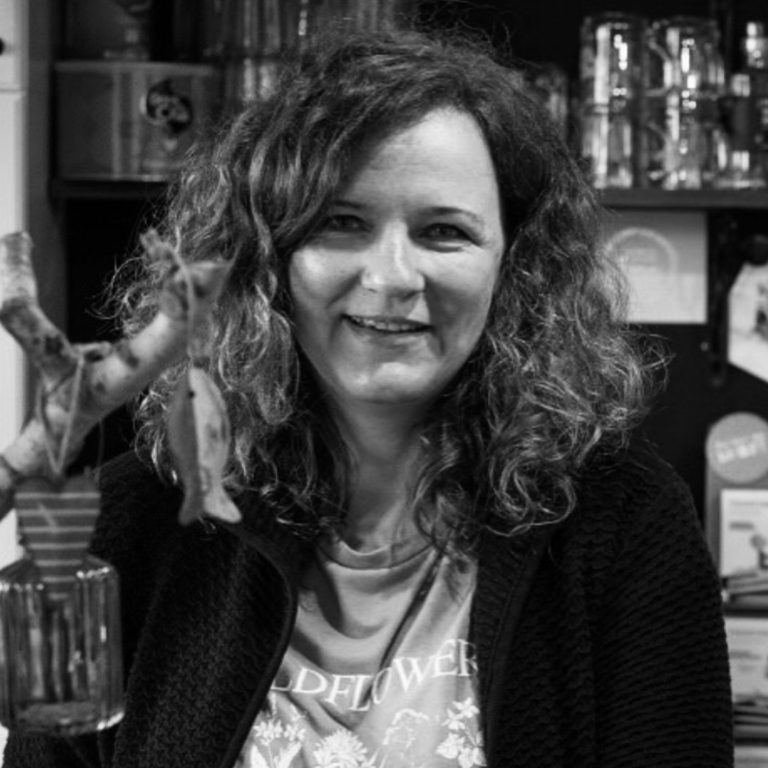 Kirstin Zöllner Frau mit lockigem Haar, lächelt vor einem Hintergrund aus Regalen und Gegenständen.