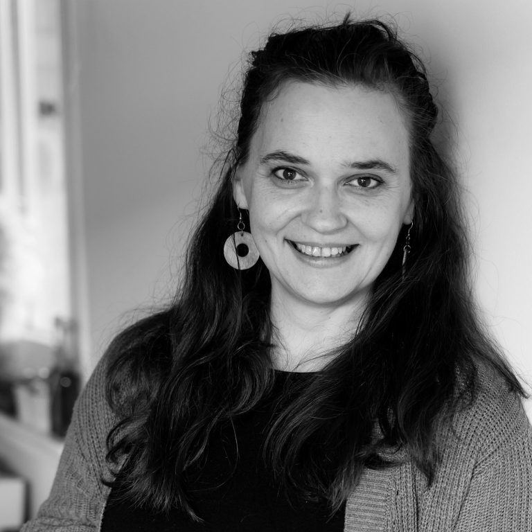 Claudia Drossert Eine lächelnde Frau mit langen, lockigen Haaren und Ohrringen, in einem hellen Raum.