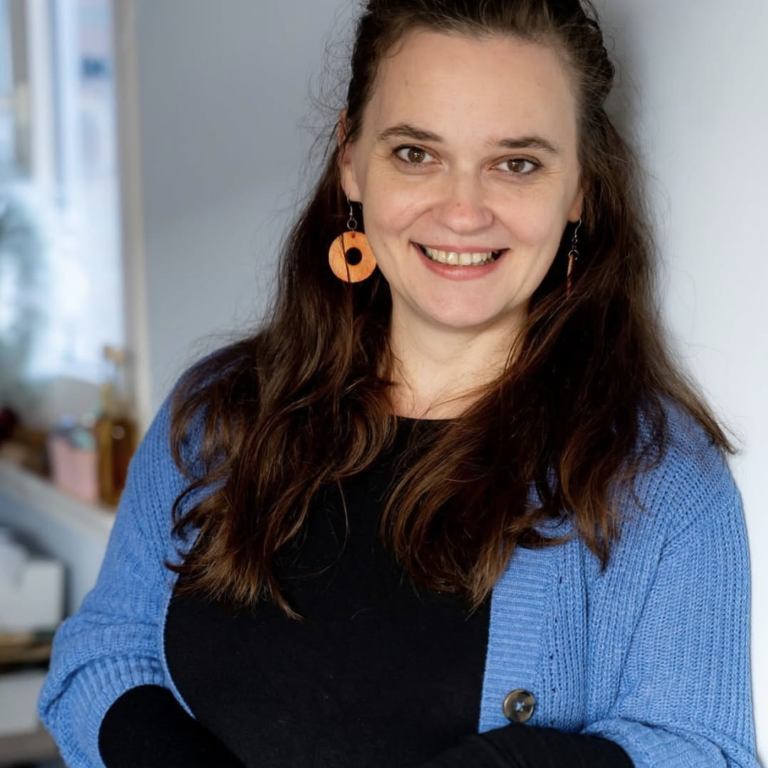 Claudia Drossert Zitat Lächelnde Frau mit langen Haaren und orangefarbenen Ohrringen, trägt einen blauen Cardigan.