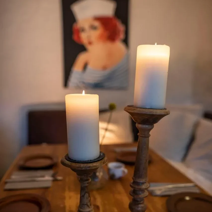 Kleine Orte erzählen die größten Geschichten Zwei Kerzen auf einem Tisch, im Hintergrund ein Bild einer Frau mit rotem Haar.
